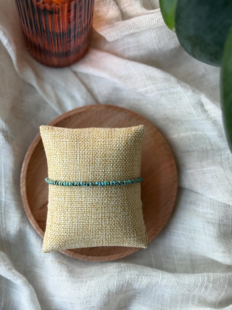 May include: A delicate turquoise beaded bracelet rests on a small, textured, yellow pillow. The pillow sits on a wooden plate, with a blurred background of a glass and greenery. The bracelet is a simple, elegant piece of jewelry.