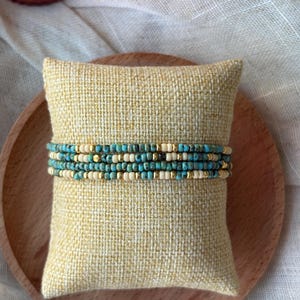 Op de afbeelding: Een set kralenarmbanden in turquoise, crème en goud. De armbanden worden gepresenteerd op een klein, vierkant, geel kussen met een getextureerde afwerking. Een houten bord en witte doek vormen de achtergrond.
