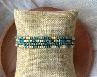 May include: Stack of beaded bracelets in turquoise, cream, and gold colors. The bracelets are displayed on a small, square, yellow pillow with a textured surface. The background includes a wooden plate and a white cloth.
