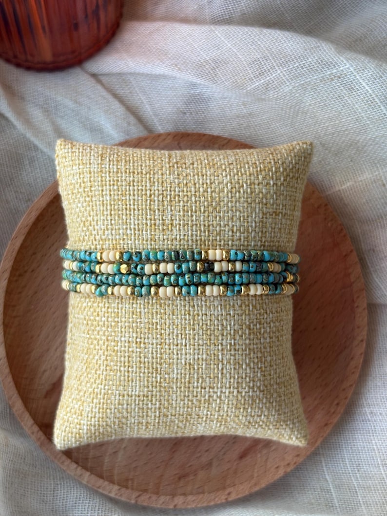 May include: Stack of beaded bracelets in turquoise, cream, and gold colors. The bracelets are displayed on a small, square, yellow pillow with a textured surface. The background includes a wooden plate and a white cloth.