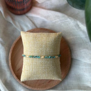 May include: A beaded bracelet with turquoise and gold-colored beads is displayed on a small, square, tan pillow. The pillow rests on a round, wooden plate. The background includes a white cloth and a decorative glass.