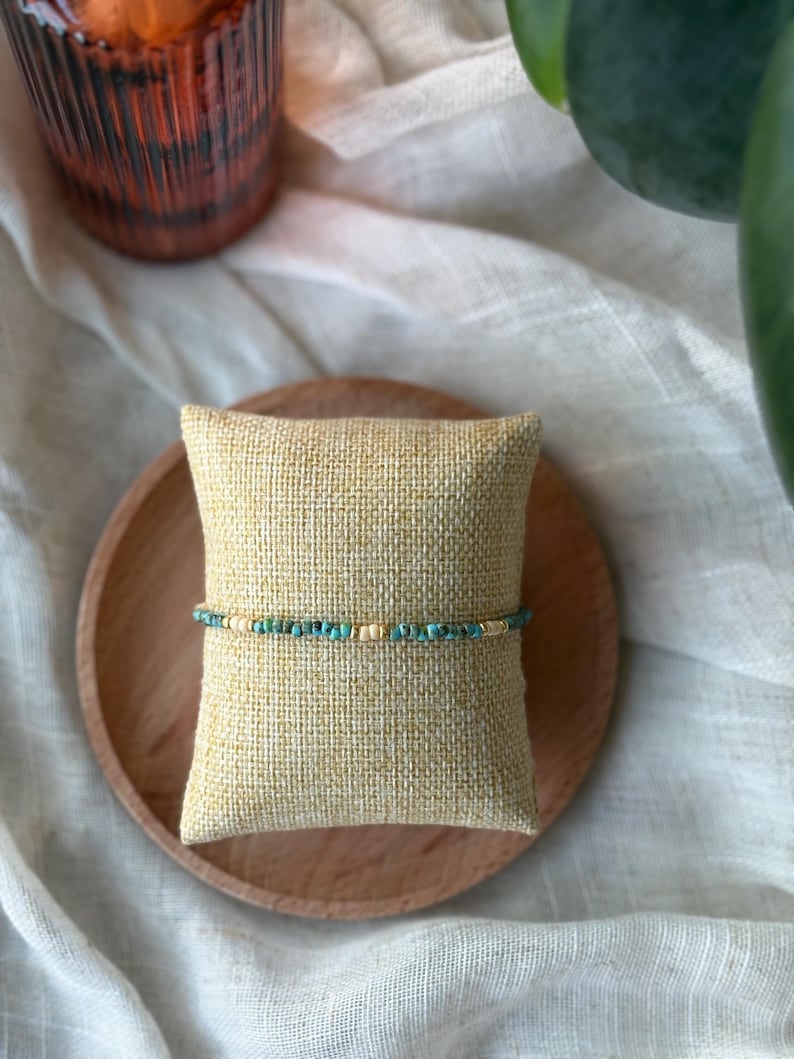 May include: A beaded bracelet with turquoise and gold-colored beads is displayed on a small, square, tan pillow. The pillow rests on a round, wooden plate. The background includes a white cloth and a decorative glass.