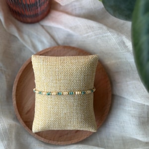 May include: A beaded bracelet with turquoise and cream-colored beads. The bracelet is displayed on a small, beige pillow, which is placed on a wooden plate. The background includes a soft, neutral-colored fabric and a green plant.