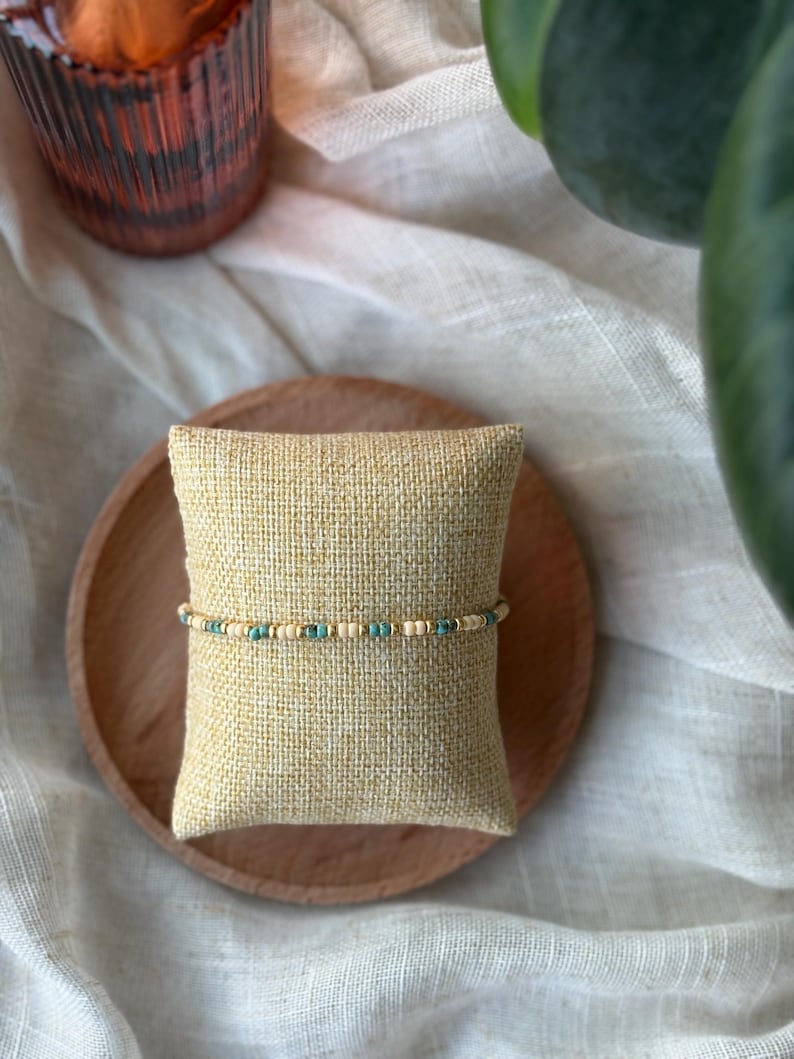 May include: A beaded bracelet with turquoise and cream-colored beads. The bracelet is displayed on a small, beige pillow, which is placed on a wooden plate. The background includes a soft, neutral-colored fabric and a green plant.