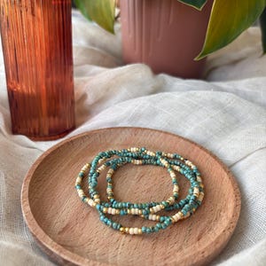 May include: A stack of beaded bracelets in turquoise, cream, and gold tones rests on a small wooden plate. The bracelets are made of small, round beads. An amber-colored glass bottle and a potted plant are in the background.