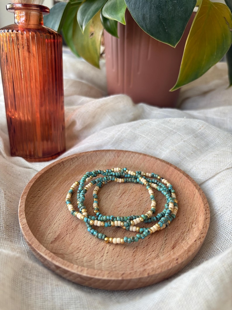 May include: A stack of beaded bracelets in turquoise, cream, and gold tones rests on a small wooden plate. The bracelets are made of small, round beads. An amber-colored glass bottle and a potted plant are in the background.