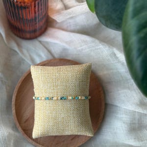 May include: A beaded bracelet with turquoise, gold, and cream-colored beads. The bracelet is displayed on a small, square, beige pillow, which rests on a round, wooden plate. The background includes a white cloth and a green plant.