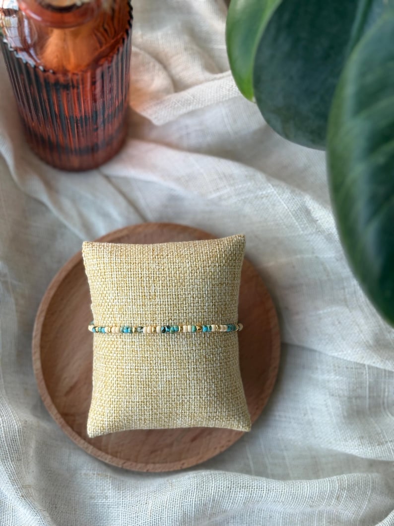 May include: A beaded bracelet with turquoise, gold, and cream-colored beads. The bracelet is displayed on a small, square, beige pillow, which rests on a round, wooden plate. The background includes a white cloth and a green plant.