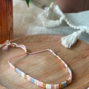 May include: A delicate bracelet made with small, square-shaped beads in shades of pink, white, and gray. The beads are separated by gold-colored spacers, creating a subtle and elegant design.