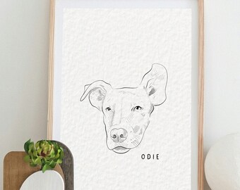 May include: A framed black and white line drawing of a dog's head with the name "ODIE" printed below the image. The frame is light wood and the drawing is on a white background. The drawing is on a white dresser with two drawers.