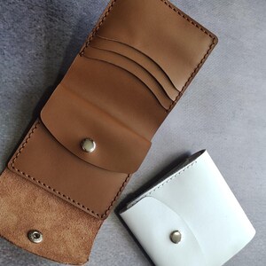 以下が含まれることがあります： 3つのカードスロットとスナップボタン留め具を備えた開いた状態のタンカラーのレザーウォレットと、同じくシルバーカラーの金属製スナップボタン留め具が付いた閉じた状態の白いレザーウォレット。どちらのアイテムも縁に目に見えるステッチが施されています。