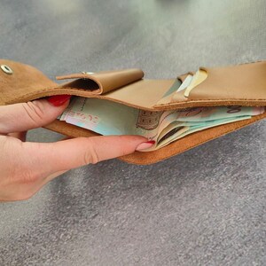 以下が含まれることがあります： スナップボタン留めのタンカラーのレザーウォレット。開いた内部コンパートメントには数枚の紙幣が入っています。このウォレットは滑らかなライトブラウンのレザーで作られており、縁には目に見えるステッチが施されています。