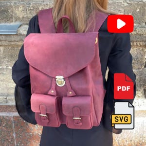 May include: A deep burgundy leather backpack featuring a large fold-over flap with a central brass-toned metal push-lock closure, two front pockets with buckle straps, a top handle, and adjustable shoulder straps for carrying on the back.