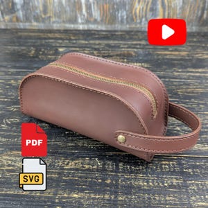 May include: A brown leather toiletry bag with a curved top, a gold-toned metal zipper closure, and a matching leather side handle secured with a single brass-colored rivet. The bag features visible stitching along the edges and a structured design.