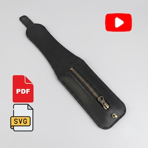 Può includere: Un bracciale in pelle nera con uno scomparto centrale dotato di cerniera in metallo color ottone e una chiusura a borchia color ottone sul fondo. Il cinturino ha una forma affusolata con un piccolo foro all'estremità più stretta.