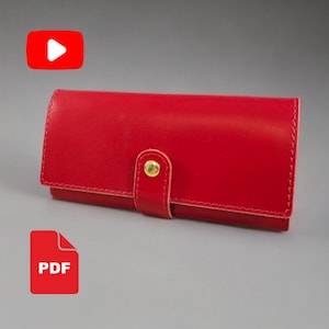 Puede incluir: Una cartera rectangular de cuero rojo con una solapa plegable asegurada por un botón de presión de metal dorado. La cartera presenta costuras visibles a lo largo de los bordes y una lengüeta de cierre central con un acabado liso.