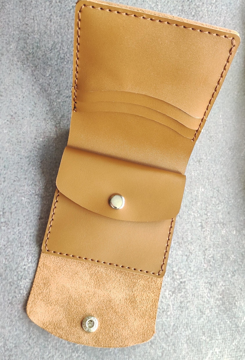 以下が含まれることがあります： 滑らかな仕上げのタンカラーのレザー製二つ折り財布。縁には目に見えるステッチが施され、中央にはシルバーカラーの金属製スナップボタン付きコインポケットがあります。内側には複数のカードスロットと、もう一つのスナップボタン留め具が備わっています。