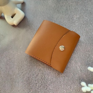 以下が含まれることがあります： 中央にシルバーカラーの金属製スナップボタンが付いた、正方形のタンカラーのレザーウォレット。フラップの中央に留め具があり、下端に沿って目に見えるステッチが施されています。全体的に滑らかでマットな質感の仕上がりです。