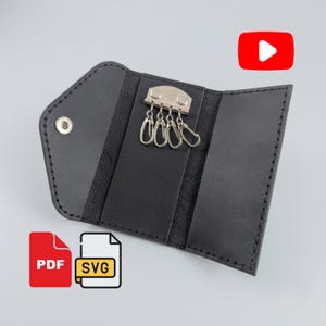 Peut inclure: Un étui à clés en cuir noir doté d'une barrette métallique argentée avec quatre mousquetons à ressort pour fixer les clés. L'étui possède un rabat avec un bouton-pression en métal argenté et des coutures noires apparentes sur les bords.
