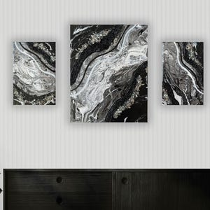 Könnte beinhalten: Ein Set aus drei abstrakten Kunstwerken mit einem marmorierten Design in Schwarz, Weiß und Silber. Die Kunstwerke werden an einer weißen Wand über einem schwarzen Schrank ausgestellt. Die Stücke variieren in Größe und Form und schaffen eine moderne Ästhetik.