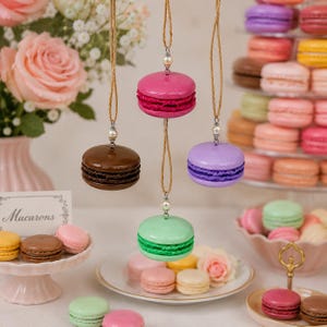 May include: A display of colorful macaron ornaments, including pink, purple, green, and brown, each suspended by a string. The macarons are arranged with a variety of real macarons and flowers, creating a festive and decorative scene.