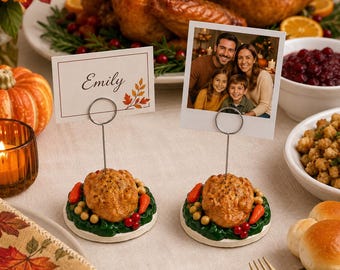 May include: Two place card holders featuring a sculpted roasted turkey surrounded by green garnish, orange carrots, and red berries on a round base. A thin silver wire extends upward from each turkey to hold a rectangular card or photo display.