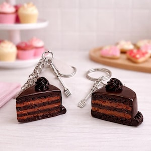 以下が含まれることがあります： チョコレートケーキのカットを模したキーホルダー2つ。ダークブラウンの光沢仕上げで、層状のスポンジの質感と上に乗ったクリームが特徴です。各キーホルダーには銀色の金属製リングと、小さなフォーク型のチャームが付いています。