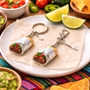 May include: Two miniature burrito keyrings with silver foil wrapping, a brown filling, and red and green toppings, sit on a white plate. Each keyring has a silver fork charm and a silver ring or clasp. The plate is surrounded by food items.