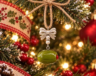 Può includere: Un addobbo per albero di Natale a forma di oliva verde con un centro rosso. L'oliva pende da un fiocco in metallo argentato con due piccole perline bianche, sospesa da un cordino rustico marrone annodato a fiocco decorativo.
