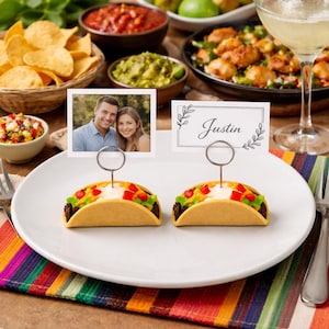 May include: Two taco-shaped place card holders made of tan material, each featuring a silver wire loop stand. One holder displays a rectangular photo of a couple, and the other holds a white card with the name Justin in black script.