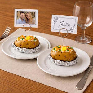 Puede incluir: Dos patatas al horno en platos blancos, cada una envuelta en papel de aluminio y cubierta con queso y verduras. Una patata tiene una foto, la otra una tarjeta con el nombre "Justin".