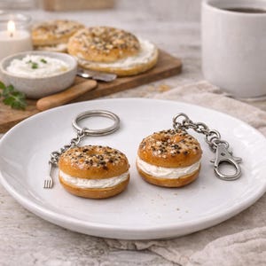 May include: Two bagel-shaped keychains with cream cheese filling sit on a white plate. The bagels have everything seasoning. Each keychain has a silver chain and clasp. A small fork charm hangs from one keychain.
