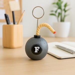 May include: A novelty desk accessory shaped like a bomb, featuring a gray sphere with a black and white "F" on the front. A twisted yellow fuse with a red tip extends from the top, and a silver metal ring is attached. The item is on a wooden desk.