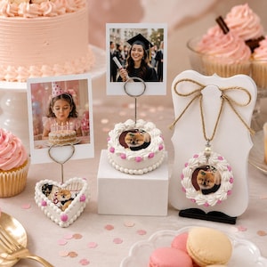 May include: Three white cake-shaped photo holders with pink frosting accents. One heart-shaped base holds a photo of a child, one round base holds a photo of a dog, and one hanging ornament features a dog photo with a jute twine bow on a white stand.