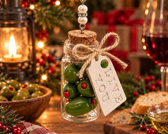 Può includere: Un barattolo di vetro trasparente riempito con olive verdi con peperoncino rosso, sigillato con un tappo di sughero. Un fiocco di spago di iuta fissa un'etichetta rettangolare con la scritta "LIVE LOVE" e un'icona di oliva verde. Il barattolo pende da un filo decorato con due perle bianche e una fascia centrale di strass.