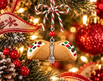 May include: A cannoli-shaped Christmas ornament featuring a golden-brown pastry shell filled with white cream and topped with red and green sprinkles. A small silver fork charm hangs from the center, suspended by a red, white, and green twine bow.