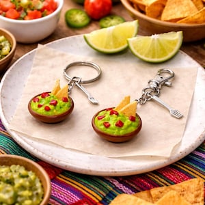 May include: Two miniature guacamole bowl keychains, each featuring a brown bowl filled with green avocado dip, red tomato pieces, and two yellow tortilla chips. Each keychain includes a silver metal ring and a small fork-shaped charm attached.