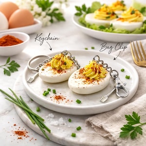 May include: Two deviled egg replicas, one attached to a metal keychain ring and the other to a metal bag clip. Each features a white egg white base, a yellow piped yolk center, and a dusting of red paprika. Both include a small fork charm.