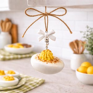 May include: A hanging ornament shaped like a deviled egg with a yellow yolk center and a dusting of paprika. The egg is suspended by a brown twine bow, featuring a white decorative bow accent and small silver-toned beads above the egg.
