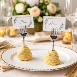 Puede incluir: Dos portatarjetas con tenedores que sostienen tarjetas con los nombres "Ethan" y "Emma". Los soportes son amarillos, en forma de espiral, y están sobre un plato blanco con borde dorado. Flores y cristalería en el fondo.