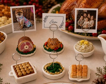 May include: A set of six miniature food-themed place card holders with silver wire clips. The collection includes replicas of a ham, a roasted chicken, mashed potatoes, a casserole, green beans, and bread rolls, each on a small base.