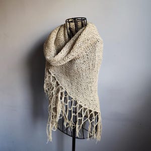 May include: A beige knitted shawl with fringe detail, draped over a wire mannequin. The shawl has a loose, open weave and a textured appearance. The fringe hangs along the bottom edge of the shawl.