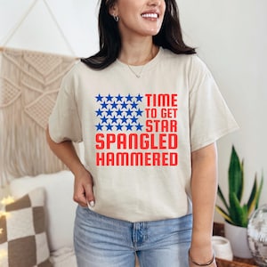 May include: A light beige t-shirt with a blue and red graphic of a star-spangled banner. The text on the graphic reads "Time to get star spangled hammered".