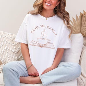 以下が含まれることがあります： 本と湯気の立つコーヒーカップの茶色の輪郭が描かれた白いTシャツ。本とカップの周りに「READ. SIP. REPEAT.」の文字が書かれている。