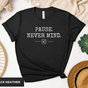 Puede incluir: Una camiseta negra jaspeada con el texto blanco PAUSE. NEVER MIND. centrado en el pecho. Debajo del texto hay un pequeño gráfico blanco de un icono de botón cuadrado con un centro circular, flanqueado por dos líneas horizontales.
