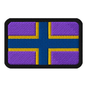 Könnte beinhalten: Gestickter Aufnäher mit einem stilisierten Flaggen-Design. Der Aufnäher hat einen schwarzen Rand, einen lila Hintergrund und ein blaues Kreuz. Goldene Akzente umranden das Kreuz und die Ränder der lila Abschnitte.