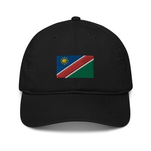 Op de afbeelding: Een zwarte baseballpet met een geborduurde vlag van Namibië op de voorkant. De vlag is blauw, rood en groen met een gele zon in de linkerbovenhoek.