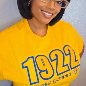 May include: A bright yellow short-sleeved t-shirt featuring the blue text 1922 and Sigma Gamma Rho printed on the front. The shirt is paired with blue denim shorts. The garment has a crew neckline and a standard fit, showcasing bold lettering.