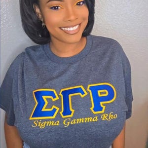 May include: A charcoal gray short-sleeve t-shirt featuring the Greek letters Sigma Gamma Rho in blue with a yellow outline, and the text Sigma Gamma Rho printed in yellow underneath. The shirt is paired with light blue denim jeans.