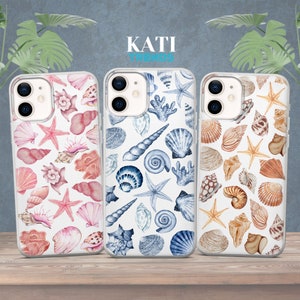 May include: Three clear phone cases with different watercolor seashell designs. The left case has pink and white seashells, the middle case has blue and white seashells, and the right case has brown and white seashells. The cases all have the text "KATI TRENDS" printed on them.